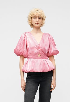 Femeie blondă cu păr creț, purtând o bluză roz strălucitoare cu mâneci bufante și detaliu răsucit în față, și pantaloni negri, pe un fundal deschis.