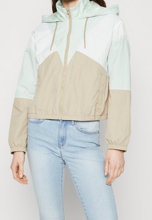 Woman wearing a light blue, beige, and white color-block zip-up hooded windbreaker with elastic cuffs and light blue jeans.