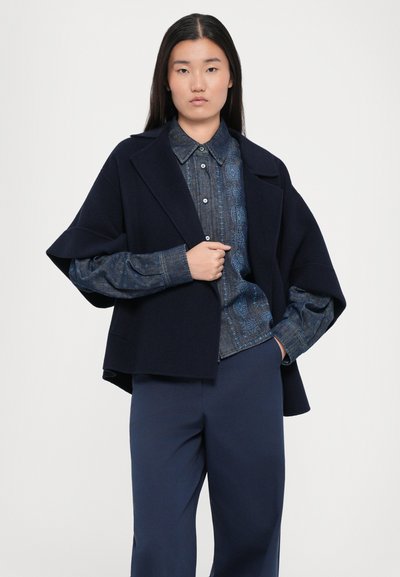 Marineblauwe wolmixjas met een brede kraag, gelaagd over een gestreept denimshirt, gecombineerd met marineblauwe broek. Gladde textuur, modern ontwerp.
