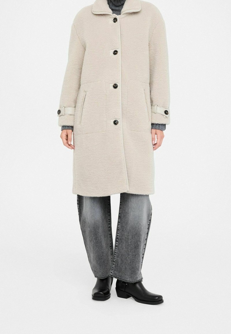 Personne portant un long manteau beige boutonné, un jean large gris délavé, des bottines noires et un pull gris en dessous.