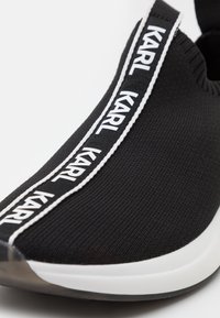 Gros plan d'une sneaker noire slip-on avec semelle blanche portant le texte « KARL » sur une bande centrale rayée noir et blanc.