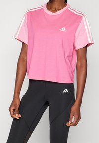 Rosa träningströja med vit tre-randig design på ärmarna och bröstlogotyp. Kortärmad, rundad hals, mjukt bomullsmaterial.