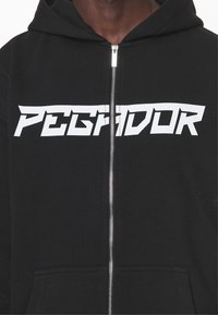 Sudadera negra con cremallera y un gran texto gráfico blanco "PEGIDOR" en el pecho. Tejido suave, bolsillo canguro y detalle de cremallera plateada.
