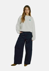 Biely oversized sveter s zeleným logom, skombinovaný s tmavomodrými širokými nohavicami z kordu a hnedými čižmami na podpätku.