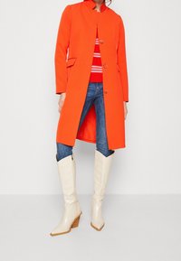 Lyse orange frakke med mandarin-krave, enkeltradet design, kombineret med en rødstribet sweater, blå jeans og hvide støvler med hæl.