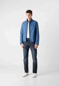 Casaco acolchoado azul com colarinho elevado, fecho de correr e padrão tonal; combinado com jeans escuros e sapatilhas brancas.