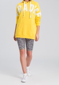 Sweat à capuche jaune avec l'inscription blanche "PARADISE", porté avec un short imprimé léopard noir et blanc et des baskets blanches.