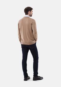 Beige Strickpullover mit V-Ausschnitt und geripptem Saum, kombiniert mit dunkelblauen Jeans und schwarzen Schuhen, von hinten betrachtet.