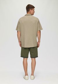 Kurzärmeliges, beiges kariertes Hemd kombiniert mit olivgrünen Shorts, die umgeschlagene Säume aufweisen, und beigen Sneakers, von hinten betrachtet.