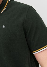 Polo shirt verde scuro con colletto a coste caratterizzato da strisce bianche e gialle. Piccolo logo bianco sul petto. Realizzato in tessuto testurizzato.