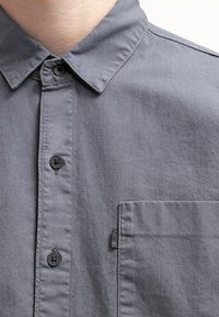 Camisa gris de manga larga fabricada en un tejido suave, con un cuello clásico, botones negros, un bolsillo en el pecho y una pequeña etiqueta de marca en el costado.