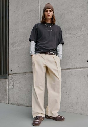 Junge Person mit brauner Mütze, schwarzem Overshirt über grauen Langarmshirts, beigen weiten Hosen und braunen Sneakers, die an einer Betonwand steht.