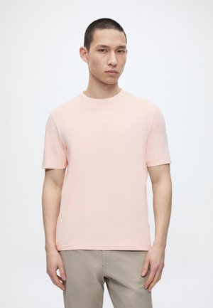 Νέος άνδρας με κοντά μαλλιά φορώντας απλό ανοιχτό ροζ κοντομάνικο μπλουζάκι και μπεζ παντελόνι, όρθιος μπροστά σε λευκό φόντο.