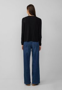 Zwarte trui met een relaxed fit, lange mouwen en geribbelde manchetten, gecombineerd met blauwe brede jeans en bruine schoenen, gezien vanuit de achterkant.