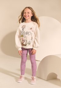 Bijela majica dugih rukava s cvjetnim i tekstualnim dizajnom, u kombinaciji s ljubičastim tajicama s otiscima plesačica. Roze tenisice zaokružuju odjevnu kombinaciju.