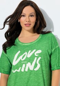 Grünes T-Shirt mit kurzen Ärmeln und rundem Ausschnitt, mit weißem Text in fetter Pinselstrich-Schrift, der "love always wins" liest.