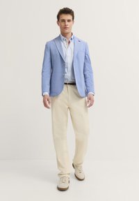 Hellblaues Sakko mit Reverskragen, hellblaues gestreiftes Hemd, cremefarbene Hose und beigefarbene Sneakers. Glattes Material und maßgeschneiderte Passform.