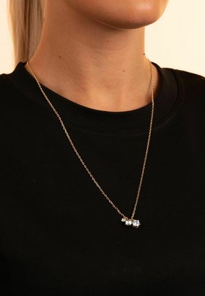 Goldfarbige Halskette mit drei klaren Kristallen an einer zarten Kette, die über einem schwarzen T-Shirt getragen wird. Die Kette besteht aus ovalen Gliedern einheitlicher Größe.