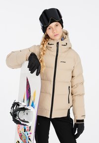 Beige gepolsterte Jacke mit Kapuze, schwarze Handschuhe und ein Snowboard mit buntem abstraktem Design. Schwarze Brille auf dem Kopf.