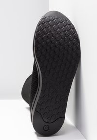 Gros plan sur la semelle en caoutchouc texturée d'une chaussure noire avec un motif hexagonal, appuyée contre un mur et un sol blancs. Aucun lacet ni détail visible.
