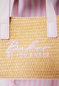 Nahaufnahme einer gewebten beigen Tasche mit hellrosa Griffen und weiß gesticktem "Baker by Ted Baker"-Logo.