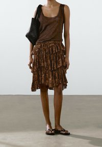 Haut marron sans manches et jupe marron superposée à motif floral. Sac fourre-tout noir tressé. Mannequin portant des tongs noires.