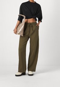 Pantalons larges verts olive en tissu texturé, associés à une chemise noire nouée et un sac beige à motifs. Le mannequin porte des chaussures à plateforme écrues.