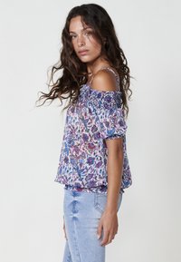 Top floreale a spalle scoperte con scollatura arricciata, caratterizzato da motivi viola, blu e rosa su un tessuto leggero, abbinato a jeans azzurri chiari.