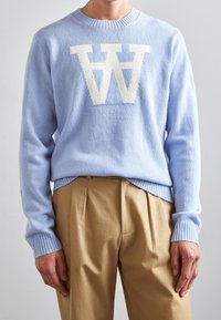 Pull en tricot bleu clair avec un logo "W" blanc très visible. Col rond et poignets côtelés avec une texture douce. Porté avec un pantalon beige.