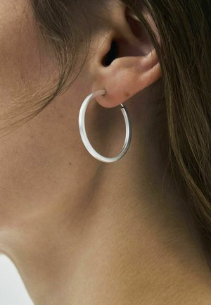 Lähikuva henkilön korvasta, jossa on sileä, keskikokoinen hopeinen rengaskorvakoru, ruskeat hiukset osittain näkyvissä.