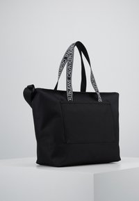 Czarna torba na ramię z przednią kieszenią i uchwytami z logo Calvin Klein Jeans w białym tekście na czarnym pasku.
