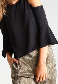 Svart off-shoulder topp med ett flödande mönster och trumpetärmar, i kombination med gyllene plisserade byxor. Slätt tyg med en mjuk textur.