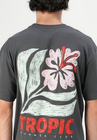 Grijze katoenen t-shirt met een groot pastel bloemenontwerp in roze en groen, met rode accenten, en vetgedrukte tekst "TROPIC SUMMER CLUB".