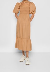 Žena v béžových midi šatech s nabíranými rukávy, smockovaným živůtkem, volánovým lemem a bílými šněrovacími robustními botami stojí před jednobarevným pozadím.