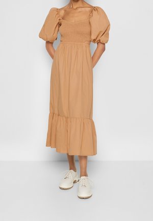 Femme portant une robe midi beige avec des manches bouffantes, un bodice froncé, un ourlet à volants et des chaussures blanches à lacets épaisses, se tenant devant un fond uni.