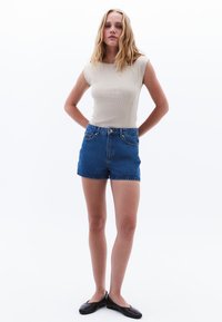 Top beige aderente senza maniche abbinato a pantaloni corti in denim blu a vita alta, con bottone frontale, cinque tasche e un aspetto leggermente consumato.