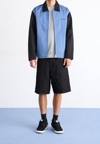 Blå og sort jakke med lynlåslukning, grå sweatshirt under, sorte shorts og sorte sneakers med hvide såler.