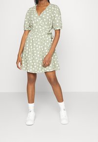 Grön prickig omlottklänning med korta puffärmar, v-ringning och knytning i midjan. Matcherad med vita sneakers och ankelsockor. Slät tygeffekt.