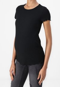 Mustassa ribattua äitiys-t-paitaa, jossa on lyhyet hihat ja aaltoileva helma. Rento malli myötäilee vartaloa, yhdistettynä tummanharmaisiin kulutettuihin farkkuihin.