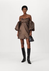 Barna selyem off-shoulder mini ruha puff ujjakkal, összehúzott mellrésszel, fekete térd fölé érő, strukturált csizmával és egy kis fekete táskával párosítva.