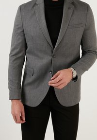 Šedý blazer s texturovaným povrchem, na dva knoflíky, s jednou náprsní kapsou, nošený přes černý rolák. Černé kalhoty a stříbrné hodinky.