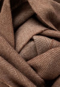 Tela marrón con patrón de espiga, que presenta una superficie suave y texturizada. El material está doblado, creando una apariencia en capas con tonos variados.