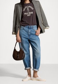 Kvinna som bär en grafisk Ralph Lauren Polo Bear-t-shirt, en grå blazer över axlarna, uppvikta blå jeans, vita sneakers och håller en brun handväska.