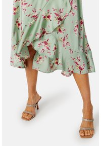 Kwiatowa sukienka w miętowym zielonym kolorze z falbaniastym dołem oraz różowo-białym wzorem kwiatowym, zestawiona z przezroczystymi sandałami na obcasie z akcentem w postaci cyrkonii.