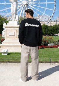 Homme portant un sweat-shirt noir avec le texte « Dreamer » dans le dos et un pantalon beige se tenant dans un parc près d'une statue et d'une grande roue sous un ciel bleu.