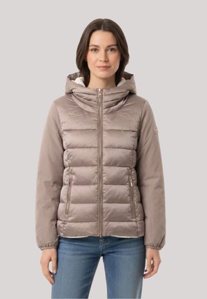 Donna che indossa una giacca imbottita con cappuccio color marrone chiaro con cerniera frontale e tasche laterali, abbinata a jeans blu, in piedi davanti a uno sfondo semplice.