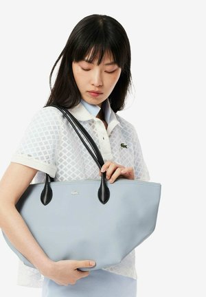 Bolso tote azul claro con asas de cuero negro. Textura suave, cierre con cremallera y un pequeño logo en el frente. Diseño clásico y espacioso.