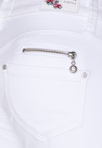Tejido de mezclilla blanca con un primer plano de un bolsillo trasero, detalle de cremallera, herrajes plateados y una etiqueta con un diseño floral.