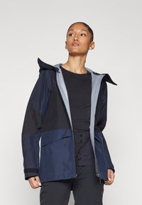 Navy- und graue wasserdichte Jacke mit Kapuze, farblich abgesetzt, mit Reißverschlusstaschen und elastischen Bündchen. Glattes, texturiertes Material.