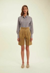 Chemise à manches longues à carreaux bleu clair et blanche, shorts plissés beiges et chaussures légères, présentés sur un fond neutre.